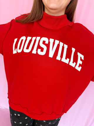 A close-up of a person wearing a bright red mock-neck sweatshirt with 'LOUISVILLE' printed across the chest in large white varsity-style letters, paired with black polka-dot pants against a pink background.