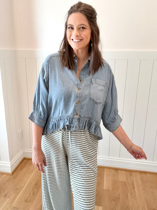 Woman wearing a denim shirt and striped pants standing in a room with wooden flooring and white walls.