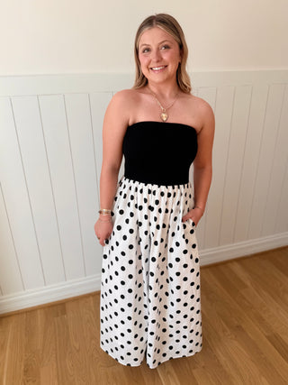 Woman wearing a black strapless top and white polka dot skirt against a white paneled wall.