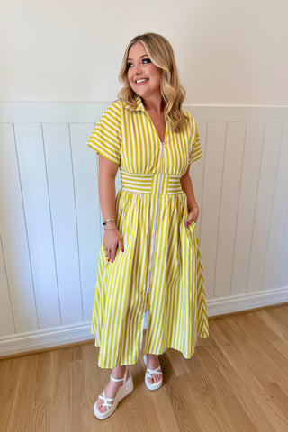 Woman wearing a yellow and white striped dress with a colorful headpiece against a white paneled wall.