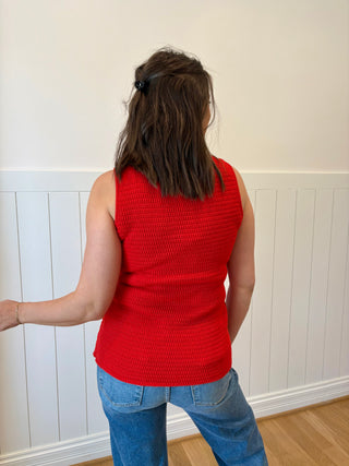 Person wearing a red sleeveless top and blue jeans standing against a white paneled wall.