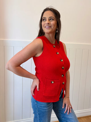 Woman wearing a red sleeveless knitted vest with buttons against a white wall.