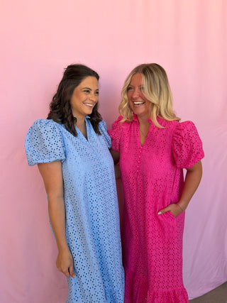 Two women wearing eyelet dresses standing against a pink background