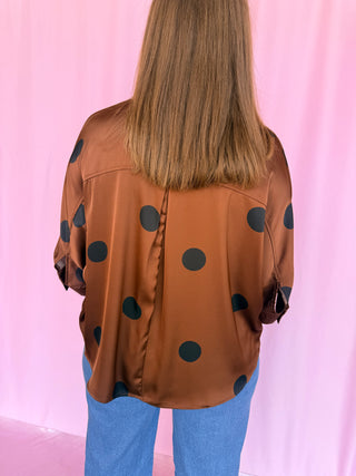 A person viewed from behind wearing a brown satin-like blouse with large black polka dots and light blue jeans, standing against a pink backdrop.