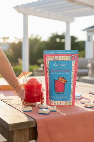 Person holding a cocktail next to a box of dominoes on a table outdoors.
