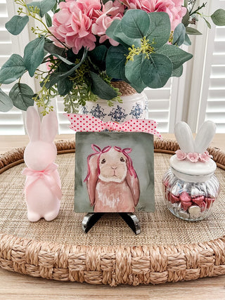 Decorative setup with a pink bunny figurine, floral arrangement, and small jar on a woven surface.