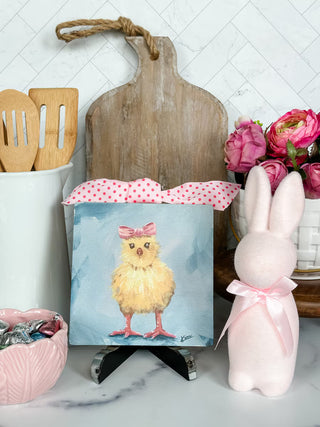 Small painting of a chick with a pink bow on a stand, surrounded by decorative items including a rabbit figurine and flowers.
