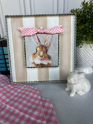 Decorative pillow with rabbit illustration and pink bow on a striped background, next to a ceramic rabbit and plant.