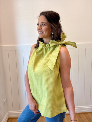 Woman wearing a yellow top with a bow detail, standing against a white wall.