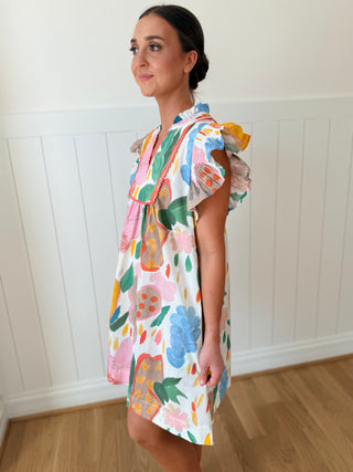 Woman wearing a colorful floral dress against a white paneled wall.