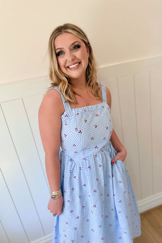 Woman wearing a blue checkered dress with a red floral headpiece against a white wall.
