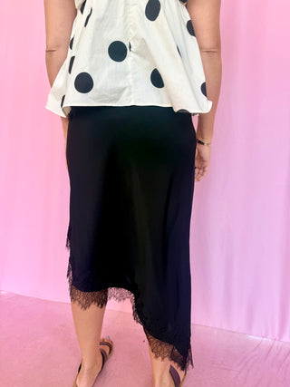 A person wearing a black midi skirt with a lace hem, paired with a cream blouse featuring gray polka dots, posed against a pink backdrop.