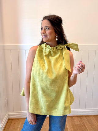 Woman wearing a yellow top with a bow detail, standing indoors against a white wall.