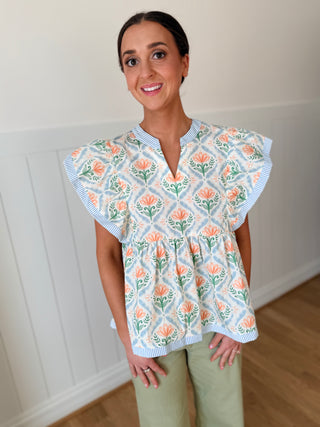Woman wearing a floral blouse with ruffled sleeves in a room with white walls and wooden flooring.