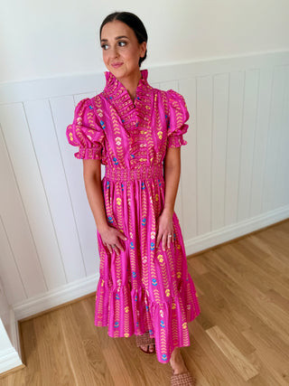 Woman wearing a pink patterned dress standing in a room with wooden flooring and white walls.