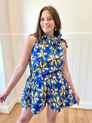Woman wearing a blue floral dress standing indoors against a white paneled wall.