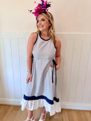 Woman wearing a white dress with blue accents and a colorful headpiece against a white paneled wall.
