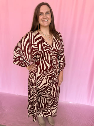 A woman wearing a brown and cream zebra-print wrap dress with three-quarter sleeves, standing against a pink backdrop.