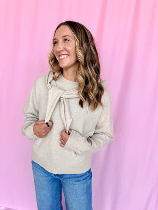 A woman wearing a light beige knit sweater with a tie-neck detail and blue jeans, standing against a pink fabric backdrop.