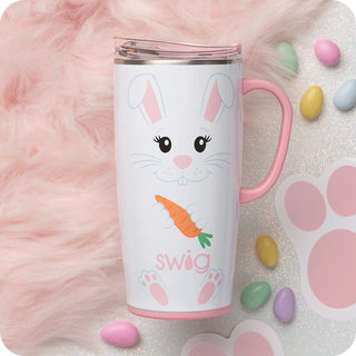 A white insulated tumbler with a pink handle and lid, decorated with a bunny face, carrot, and the word 'swig' in pink. The tumbler is styled on a glittery white surface with pastel candy pieces, a pink faux-fur texture, and a pink paw print graphic.