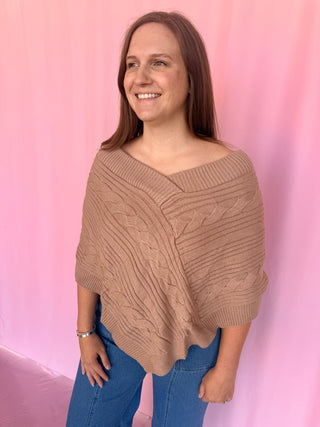A woman wearing a chunky knit off-the-shoulder poncho sweater in a warm taupe-brown color, paired with blue jeans, standing against a pink fabric backdrop.