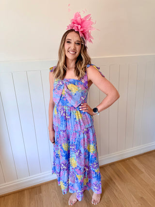 Woman wearing a colorful floral dress and pink headpiece against a white paneled wall.