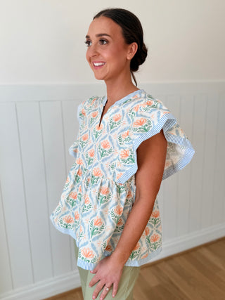 Woman wearing a floral blouse with ruffled sleeves against a white paneled wall.