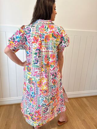 Woman wearing a colorful floral dress standing in a room with wooden flooring and white walls.