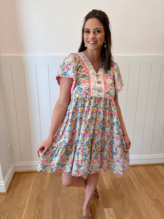 Woman wearing a floral dress standing in a room with wooden flooring and white paneled walls.