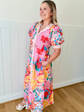 Woman wearing a colorful floral dress with a hood against a white paneled wall.