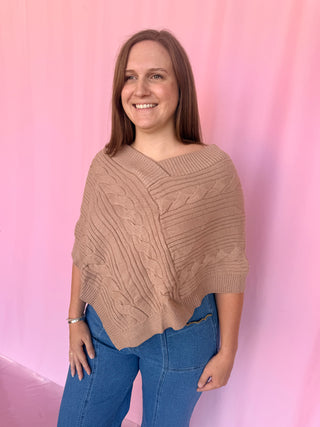 A woman wearing a beige cable-knit poncho sweater and blue jeans, standing against a pink backdrop.