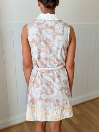 Woman wearing a sleeveless floral dress against a white paneled wall.