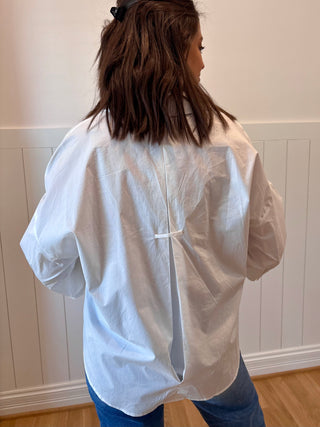 Person wearing an oversized white shirt and blue jeans against a white paneled wall.