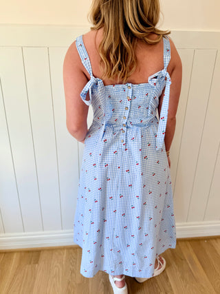Woman wearing a blue checkered dress with floral pattern against a white paneled wall.