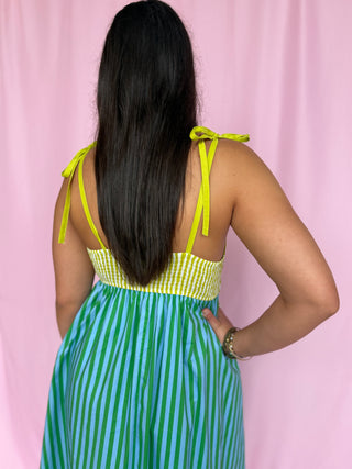 Person wearing a green and blue striped dress with yellow straps against a pink curtain background