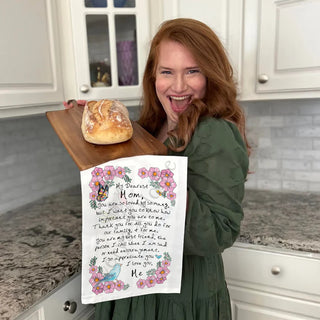 Woman holding a loaf of bread and a decorative towel with a poem in a kitchen.