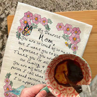 Handwritten letter to a mother with floral illustrations on a towel, held over a cup of coffee.