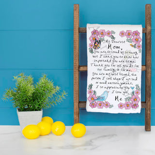 Floral-themed towel with text on a wooden ladder against a blue wall with lemons and a plant on a white surface.