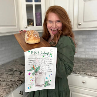 Woman holding a wooden cutting board with bread and a decorative towel in a kitchen.