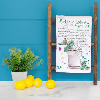 Tea towel with mint julep recipe on a wooden stand against a blue wall with lemons and a plant.