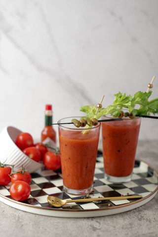 Two glasses of bloody marys with garnishes on a checkered tray with tomatoes and a bottle in the background.