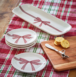 Set of white plates with red bow design on a plaid tablecloth with a wooden cutting board and butter knife.