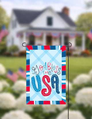 Decorative garden flag with 'God Bless the USA' text in front of a house with American flags.