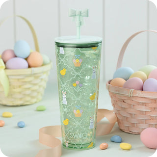 A mint green tumbler with a matching lid and bow straw, decorated with Easter-themed illustrations like bunnies, baskets, and yellow chicks. The tumbler is styled on a white surface with pastel-colored candy, ribbon, and woven baskets of eggs in the background.