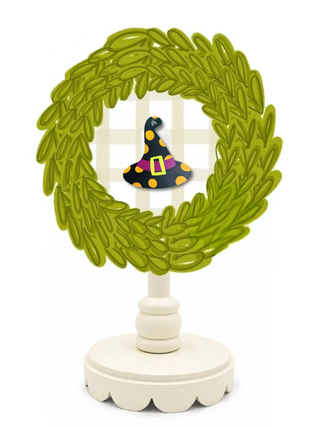 A decorative tabletop wreath with a green leafy border, mounted on an off-white pedestal stand. The center features a small black witch hat with orange and yellow polka dots and a purple band, set against a white checkered background.