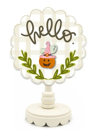A scalloped round tabletop sign on a white pedestal stand. The sign has a cream and white gingham background with the word 'hello' in dark brown script, a small orange jack-o'-lantern in a pink witch hat, a gray ghost, and green leafy branches.