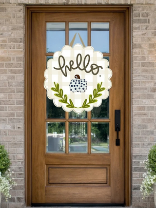 A decorative autumn-themed 'hello' door hanger is displayed on a brown wooden front door with a brick wall background. The hanger features a cream scalloped background, black cursive 'hello' text, a small blue and white polka-dot pumpkin, and green leafy branches.