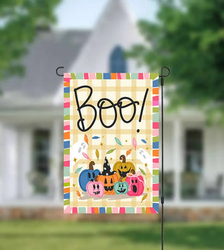 A vertical Halloween garden flag hanging from a black metal stake outdoors. The flag has a pastel multicolor striped border, a pale yellow gingham background, the word “Boo!” in large black script, and several colorful jack-o'-lanterns and ghosts near the bottom.