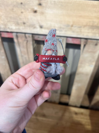 Small knitted gnome with a red name tag 'MAKAYLA' held in a hand against a wooden background.