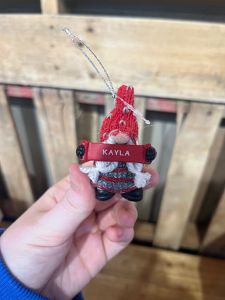 Hand holding a small gnome ornament with a 'KAYLA' tag against a wooden background
